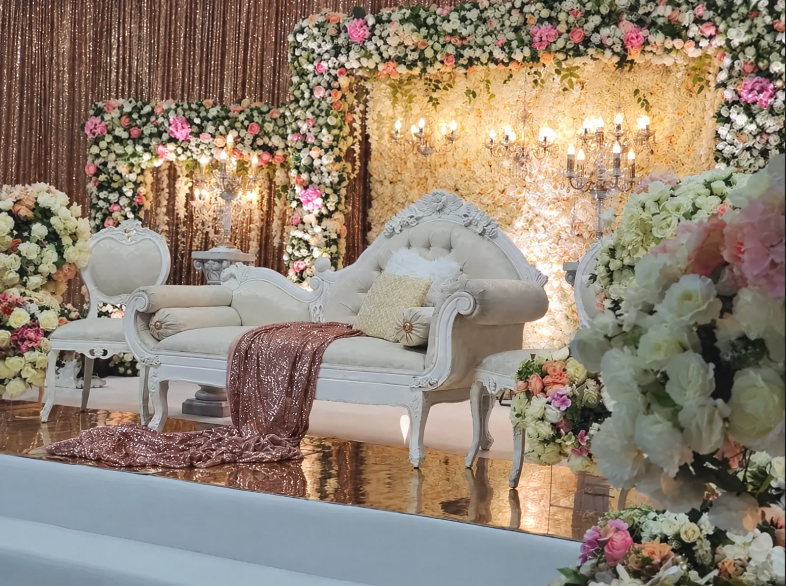 A white carved sofa with a rose gold sequin throw before blush and ivory flower walls and crystal candelabra in the Fleur 19' collection.