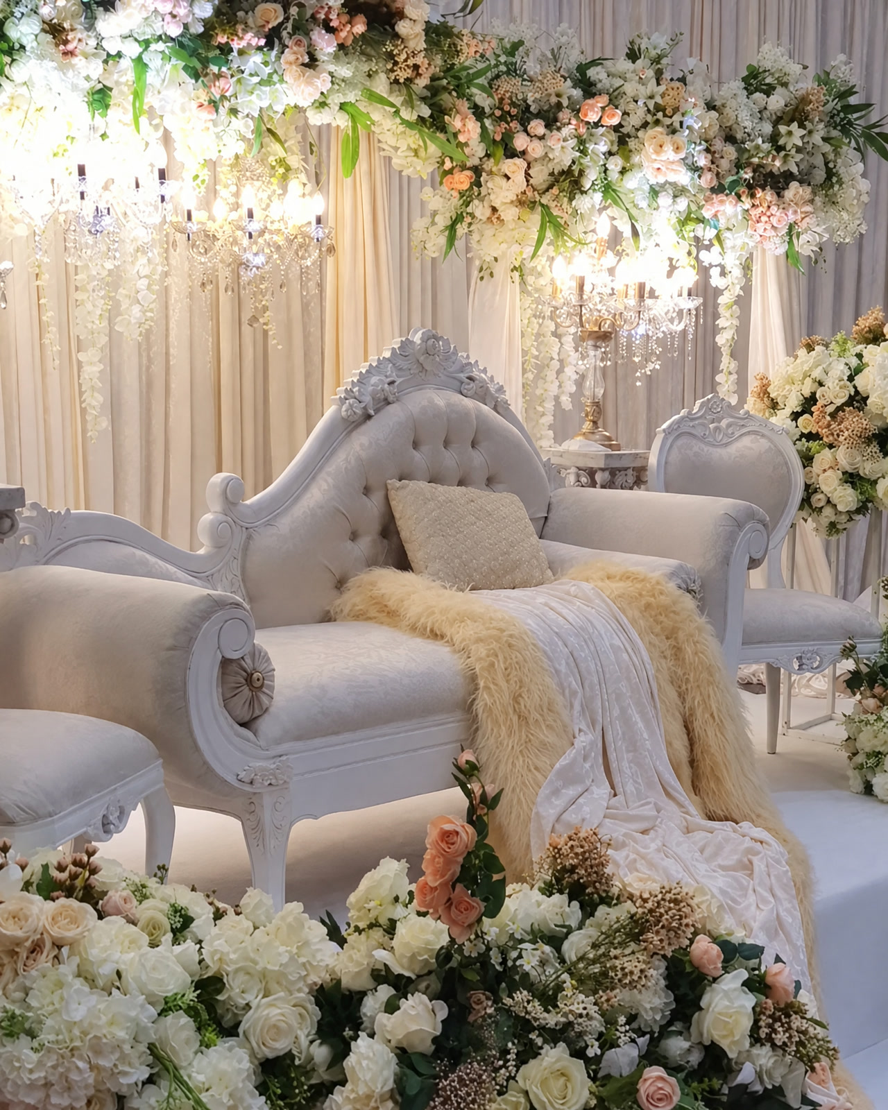 A white tufted chaise with a cream throw under cascading flowers and crystal lights, framed by ivory roses, in the Serene collection.