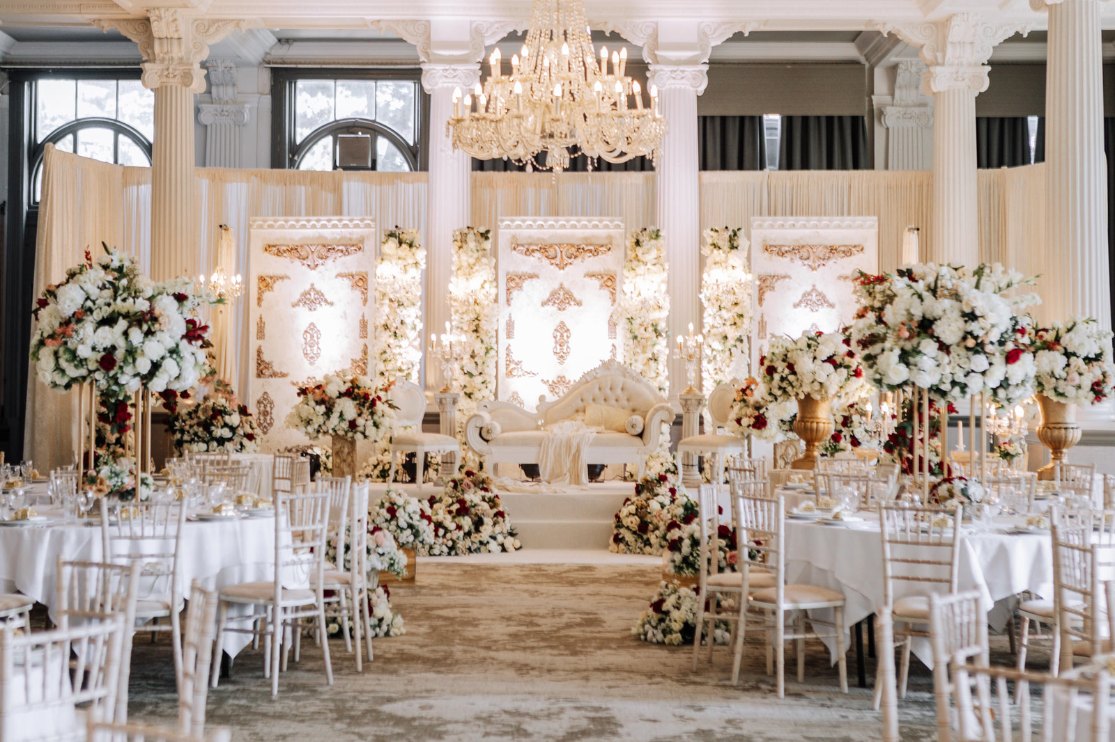 A pillared ivory hall with gilded panels, a white carved stage and tables of white and red roses in the Palace of Versailles collection.