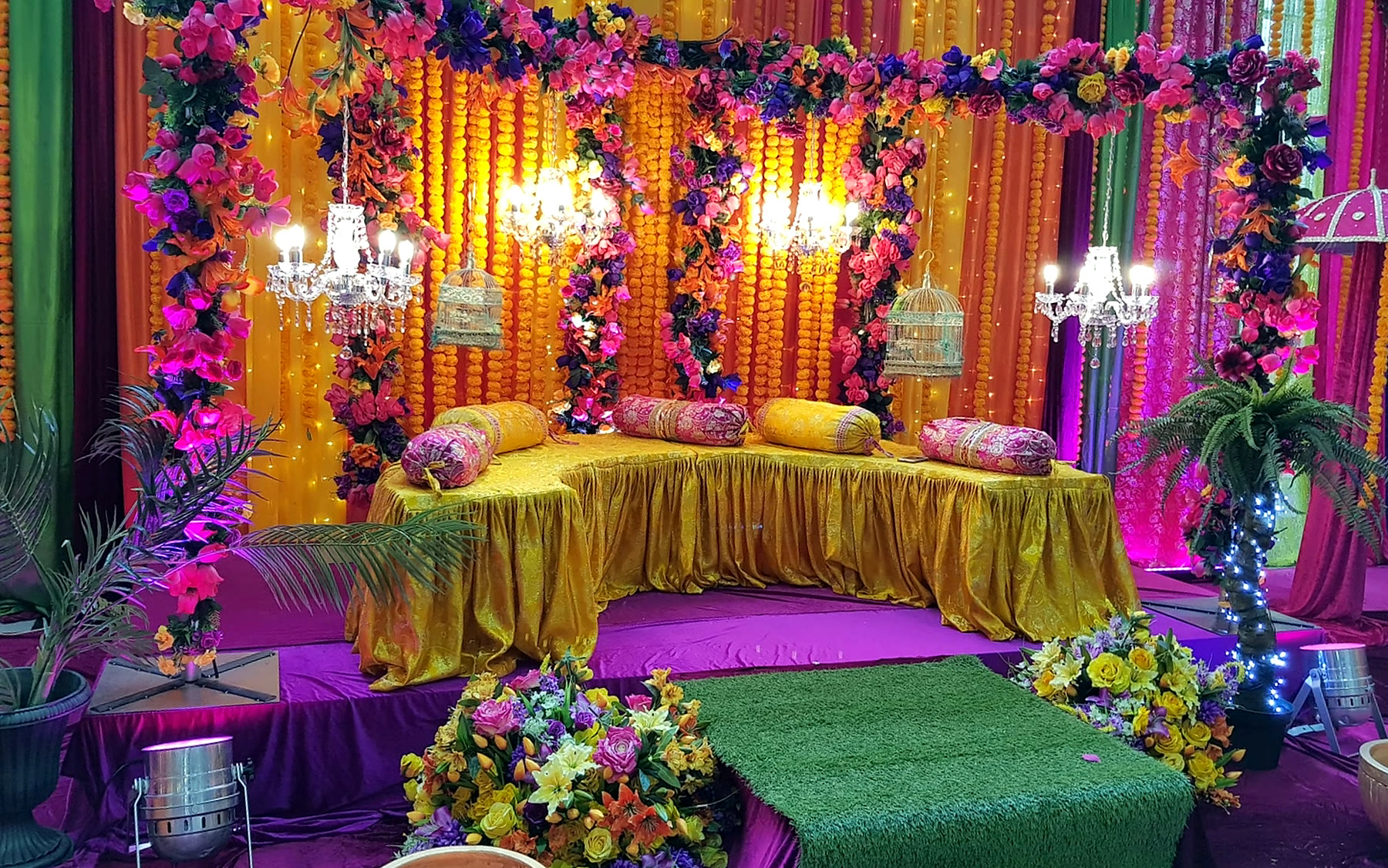 A mehndi stage in bright pink, orange and gold with flower garlands, cushions and colourful blooms.