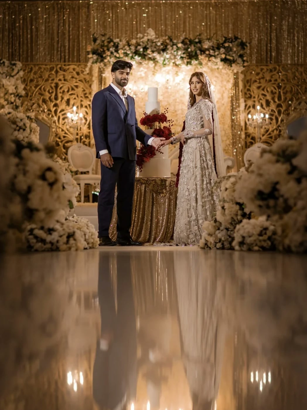 A bride and groom beside a tiered cake in front of a gold stage.