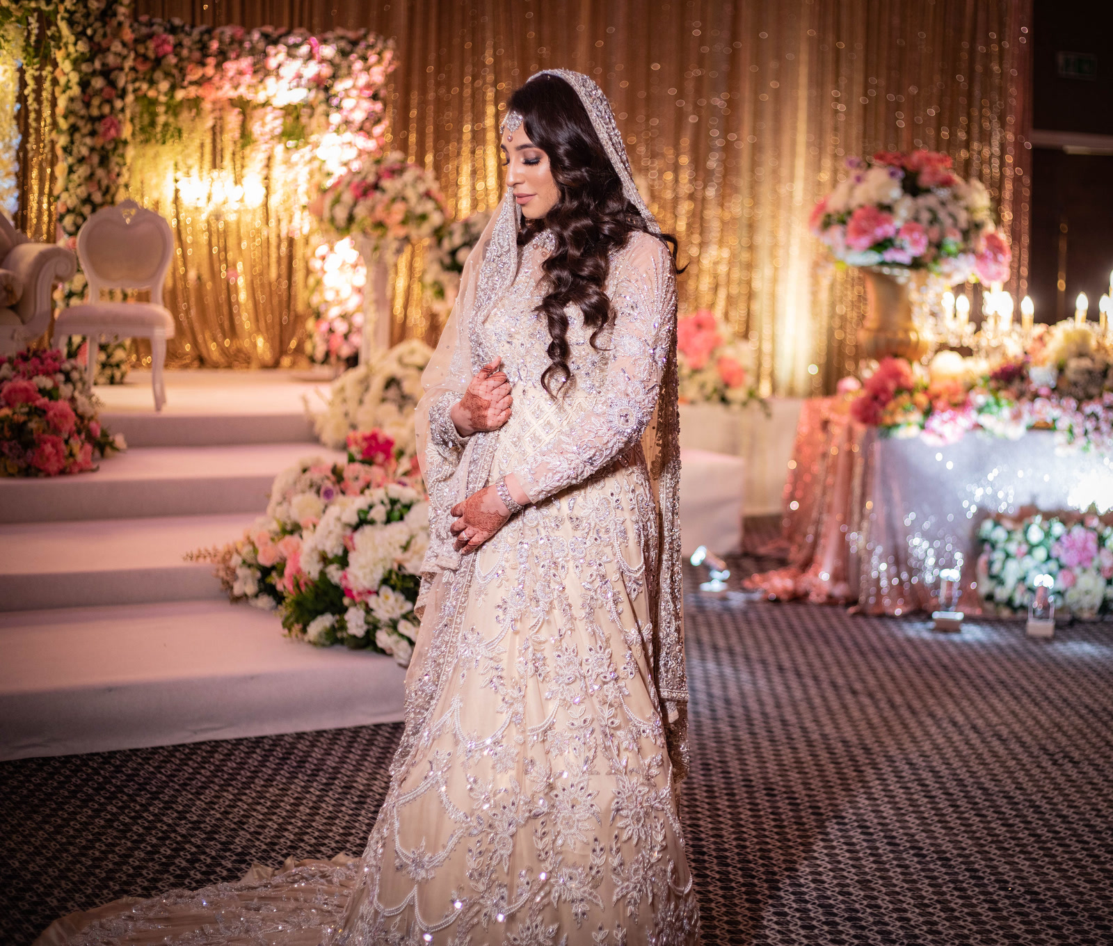 A bride in an ivory lehenga beside a stage of gold sequin and pink flowers.