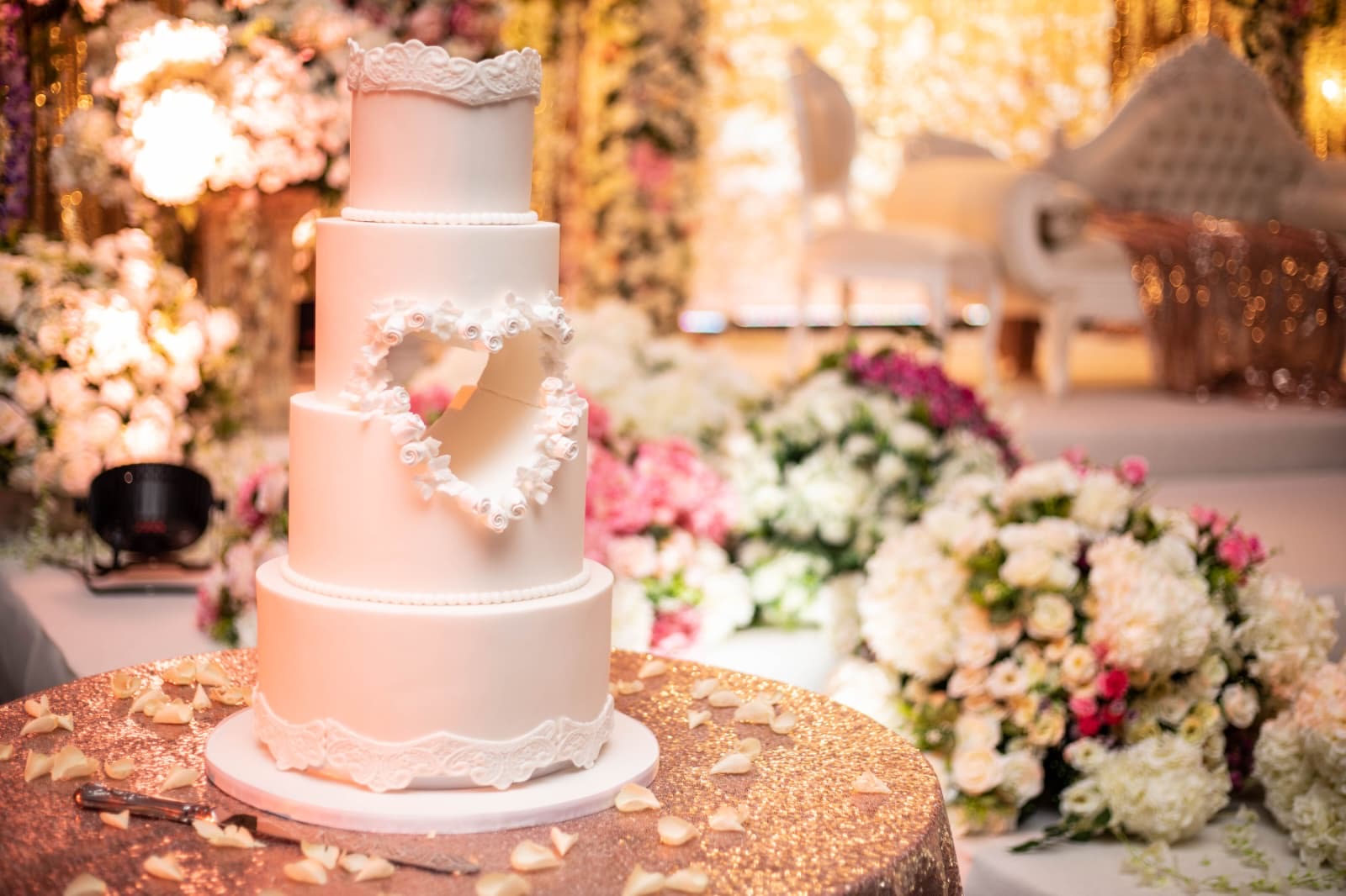 A white heart cake on a gold sequin table among pink and ivory flowers.