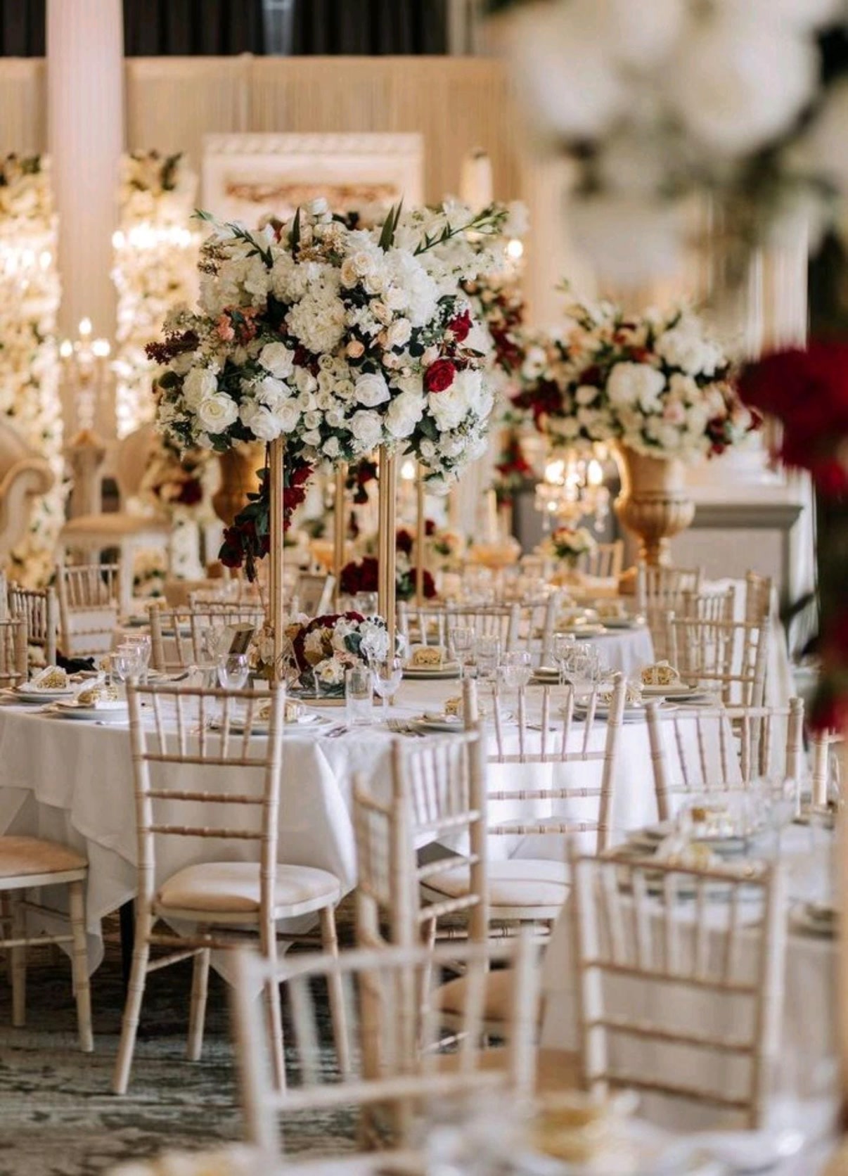 Round tables dressed with white and red flower arrangements on tall gold stands, styled by Personalize Occasionz.
